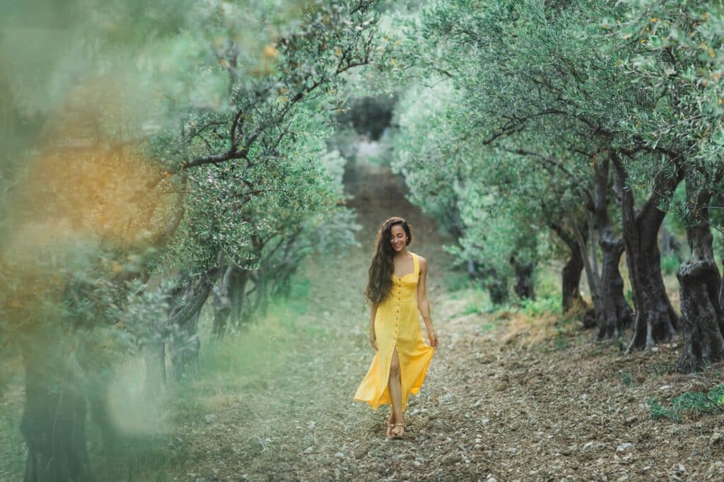 Young happy smiling woman walking in olive tree garden. Yellow linen summer dress.