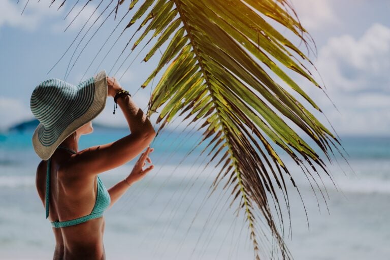 A woman with sun hat near coconut palm leaves on a tropical sandy beach. La Digue, Seychelles