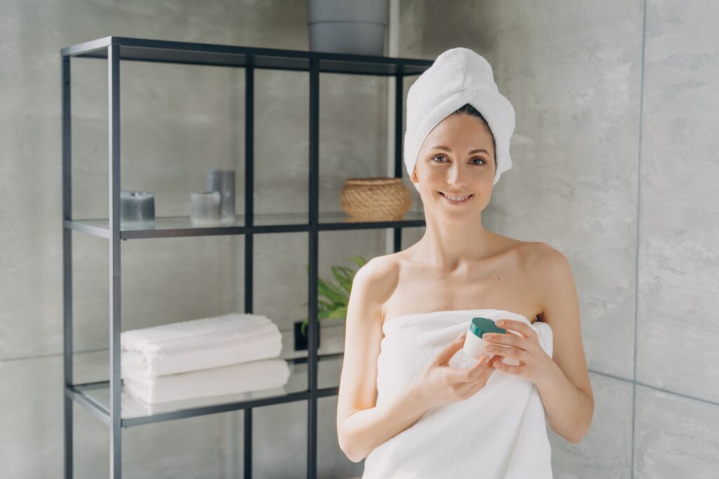 Hispanic woman holding face cream jar after shower in bathroom. Skincare daily routine, self-care